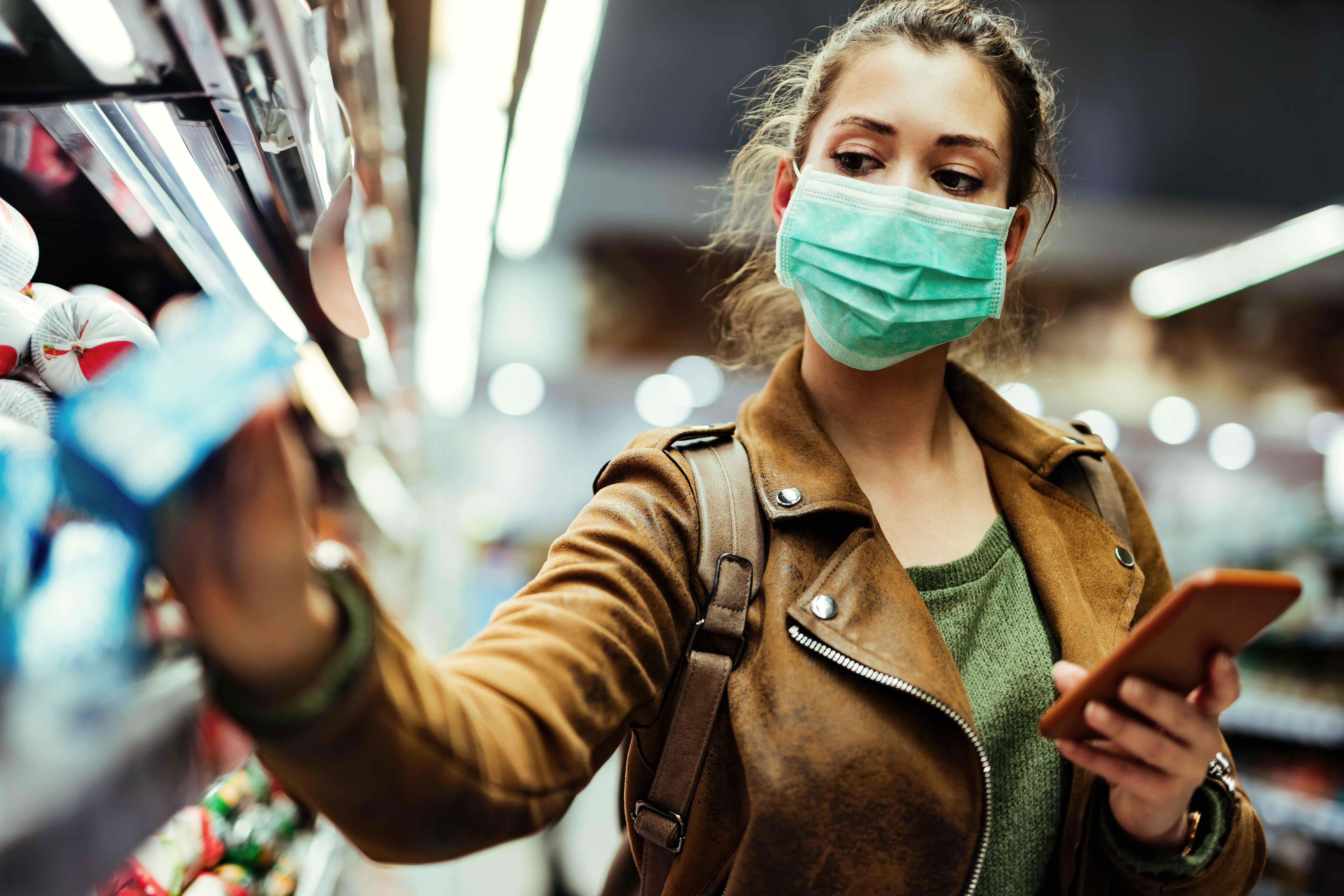 Young woman with face mask using mobile phone and buying groceries in the supermarket during virus pandemic.